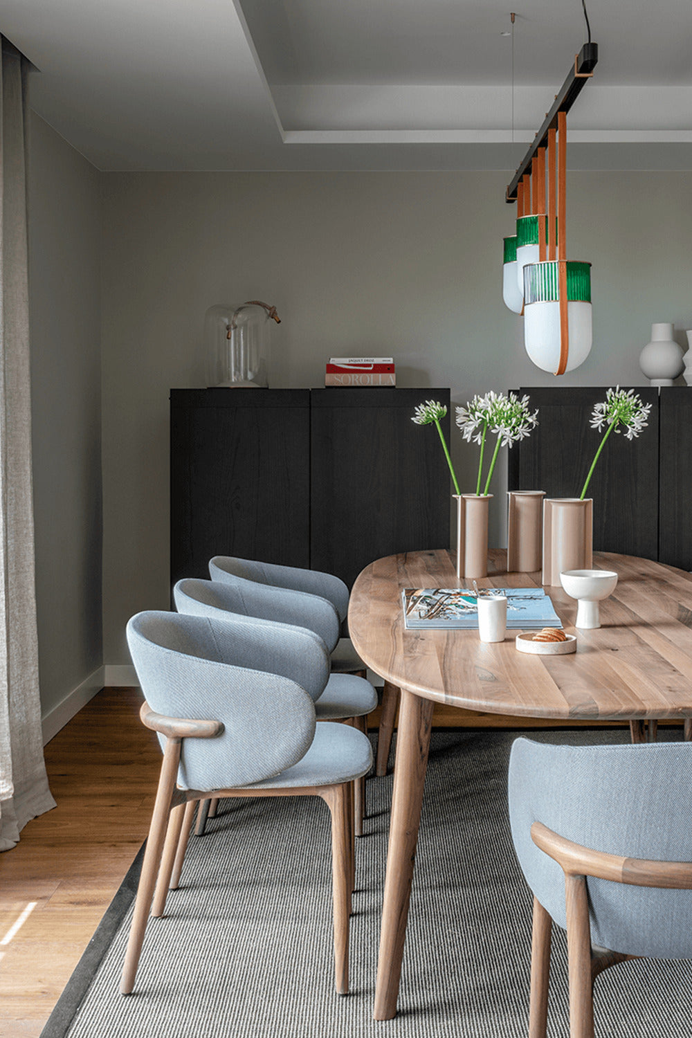 Modern dining room with wooden table, blue upholstered chairs, and stylish decor, featuring vases and hanging lights.